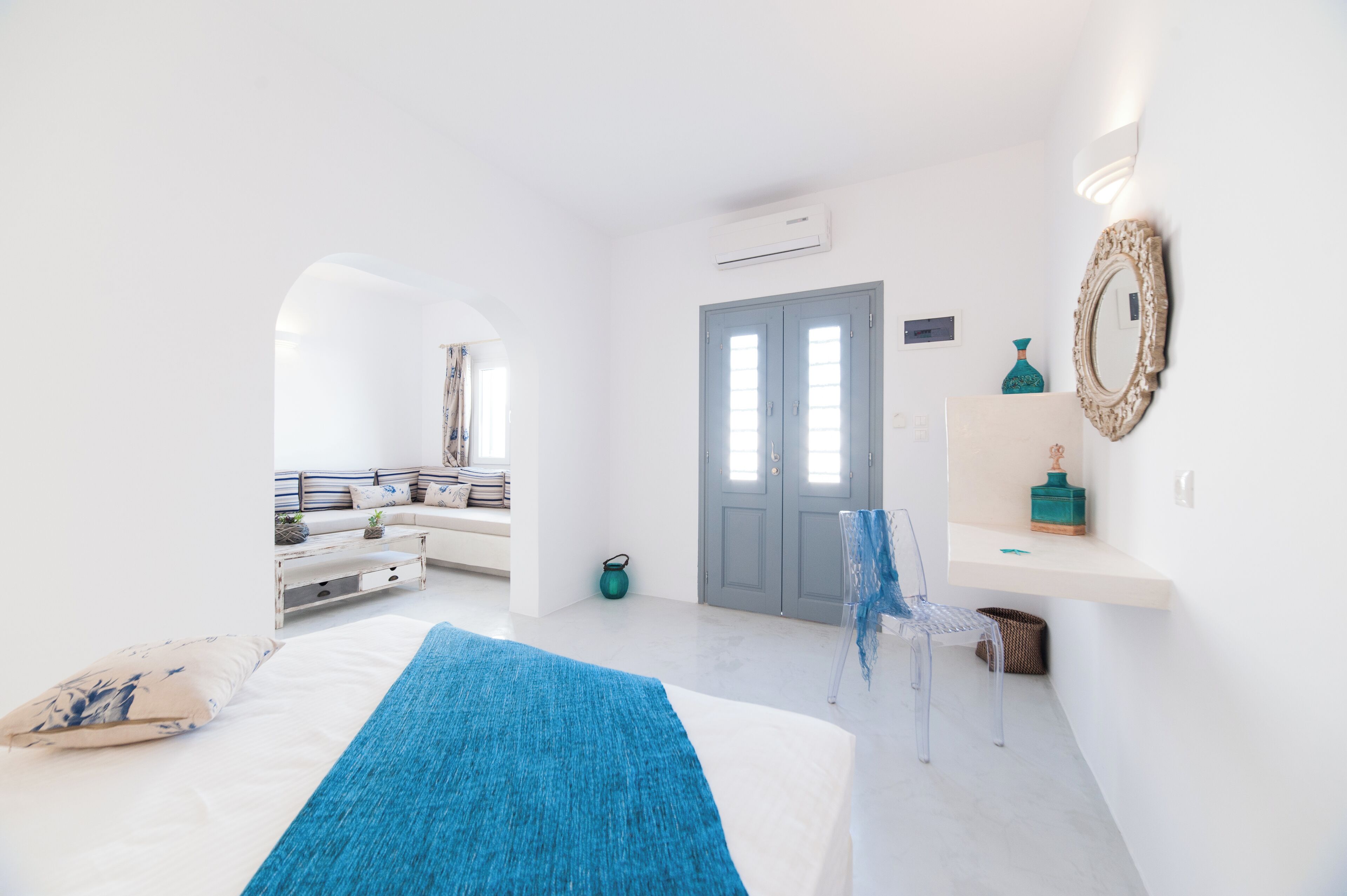 Una habitación luminosa y blanca con una cama, un tapete azul, una silla, una puerta y un estante pequeño con adornos.
