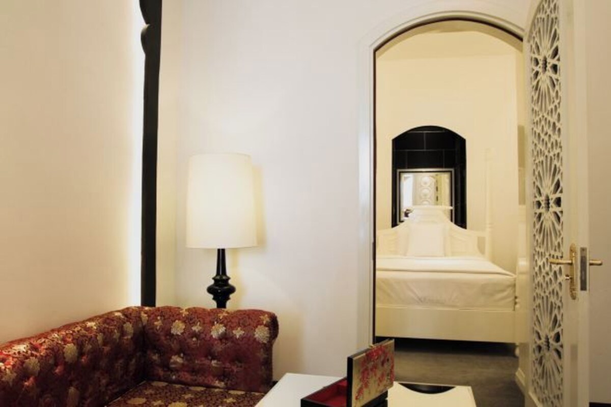 A hotel room with a floral sofa, a bedside table with a lamp, and a bed with a white headboard.