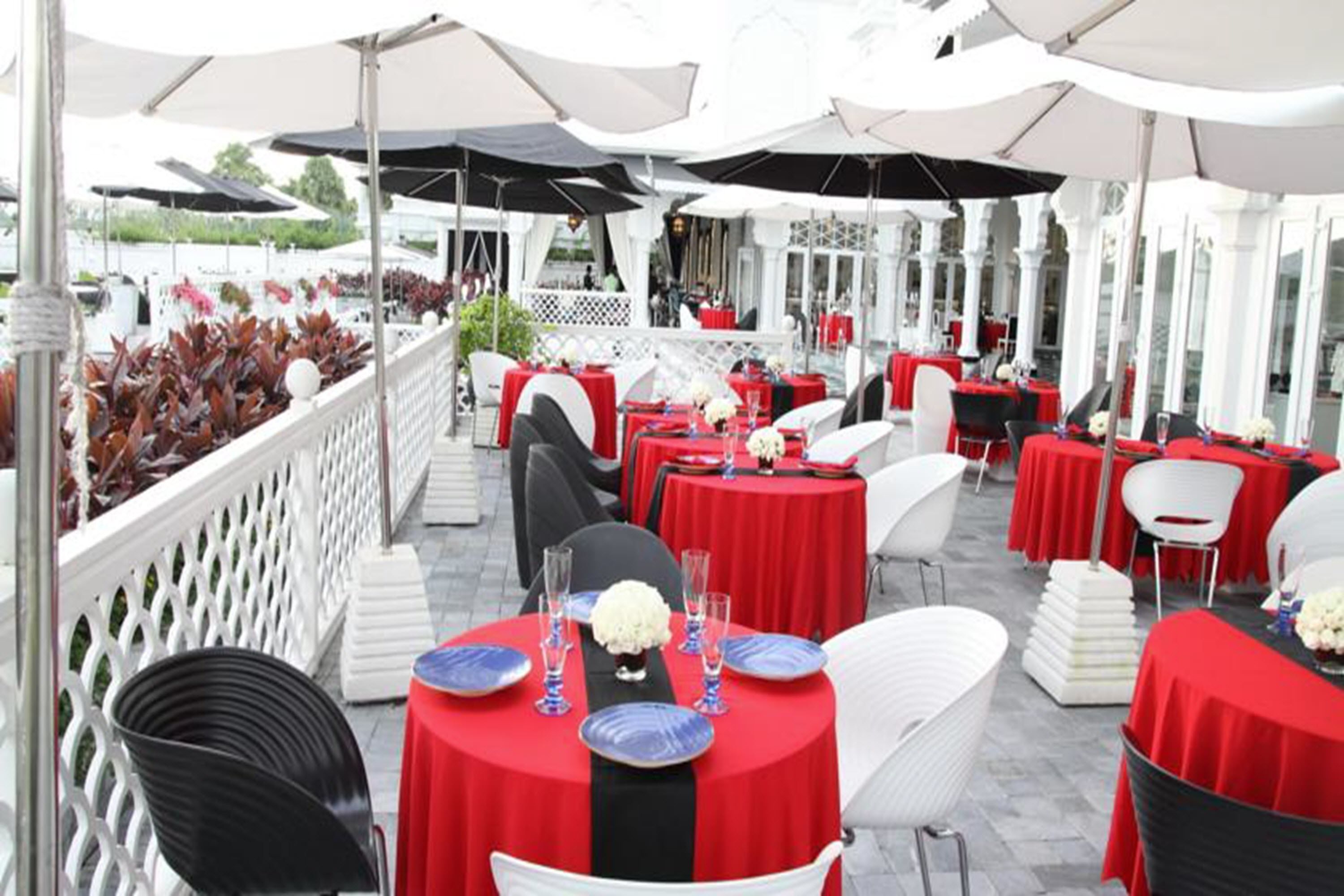 Un espacio de comedor al aire libre con mesas redondas cubiertas con manteles rojos, sillas blancas y sombrillas que dan sombra.