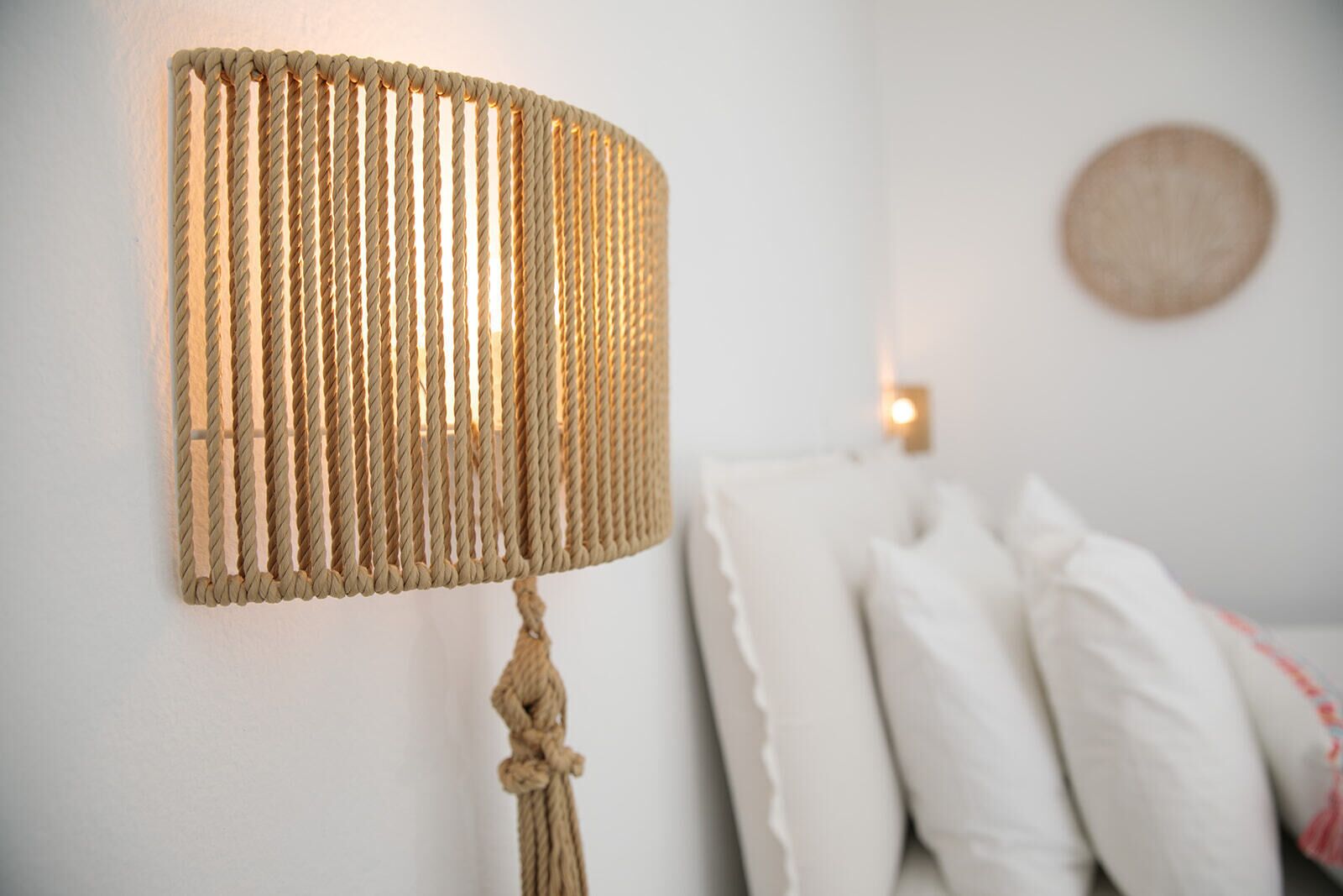 A wall-mounted lamp with a rope design, a round wall decoration, and white pillows on a bed.