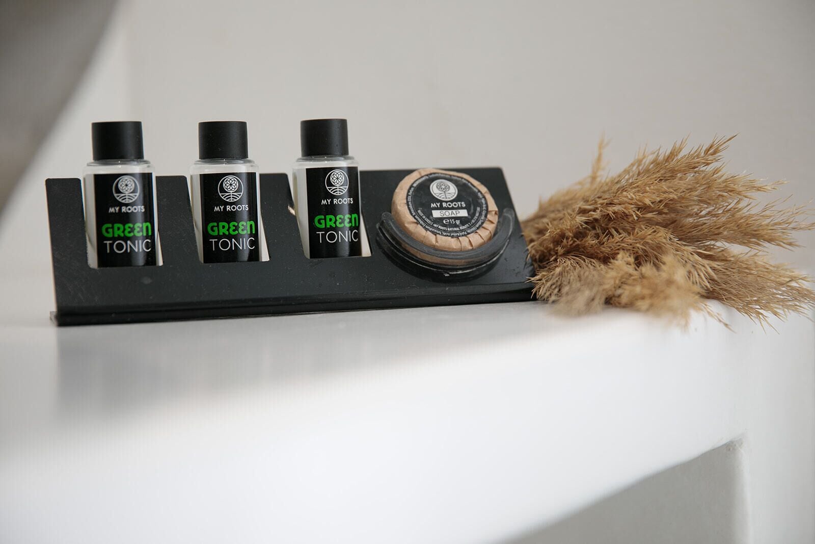 A black soap dispenser with a circular label, three bottles of green tonic, and a bundle of straw on a white surface.