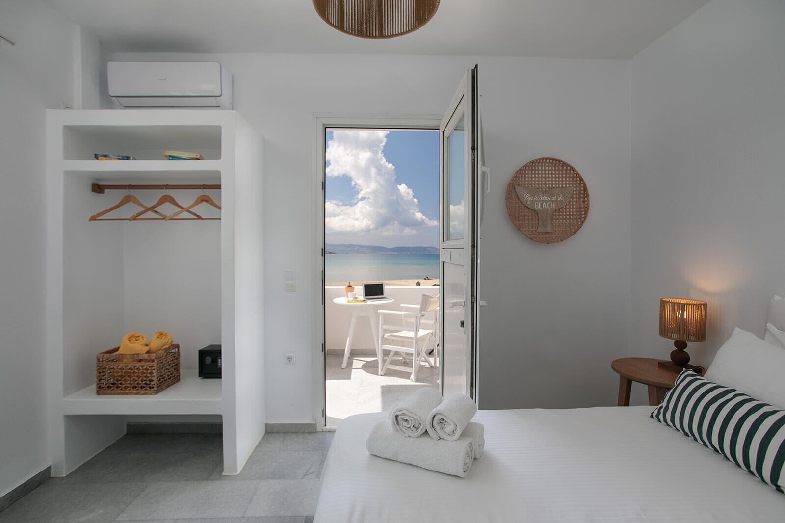 Habitación de hotel con cama, mesita de noche con lámpara, armario, pequeño balcón con mesa y sillas, y vista a la playa.