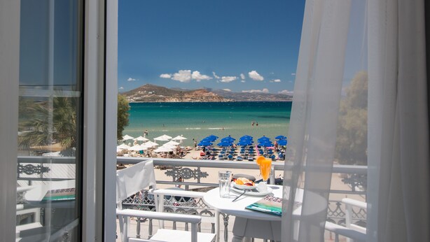 A balcony with a view of a beach, white furniture, and a table with a glass of water and a plate of food. - Ippokampos Beachfront (Naxos)