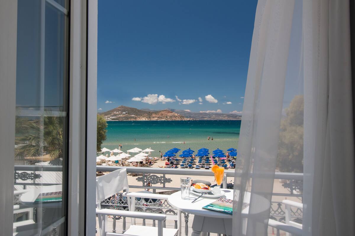 A balcony with a view of a beach, white furniture, and a table with a glass of water and a plate of food.