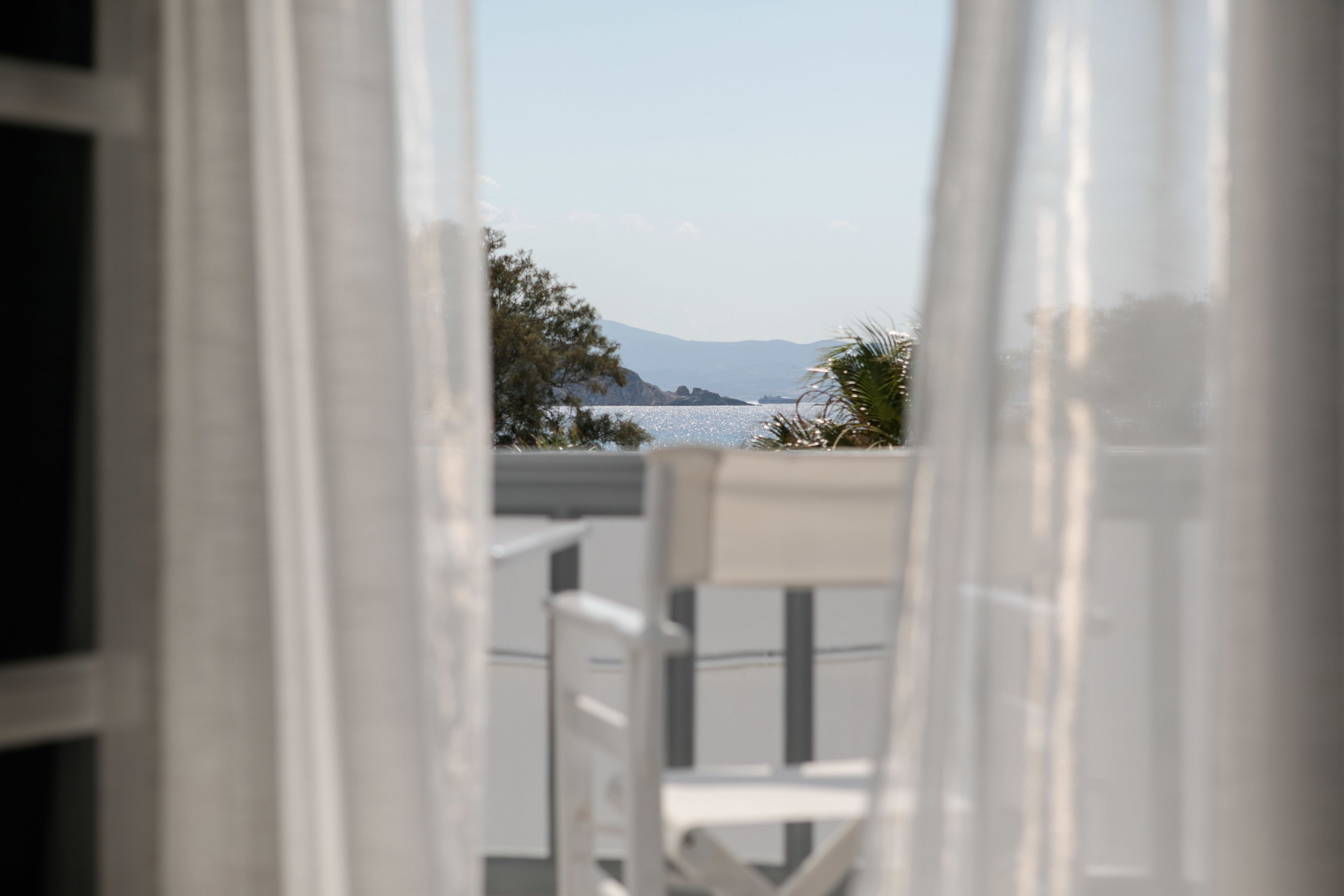 Vista por una ventana que muestra una silla blanca, un balcón y una lejana extensión de agua y montañas.