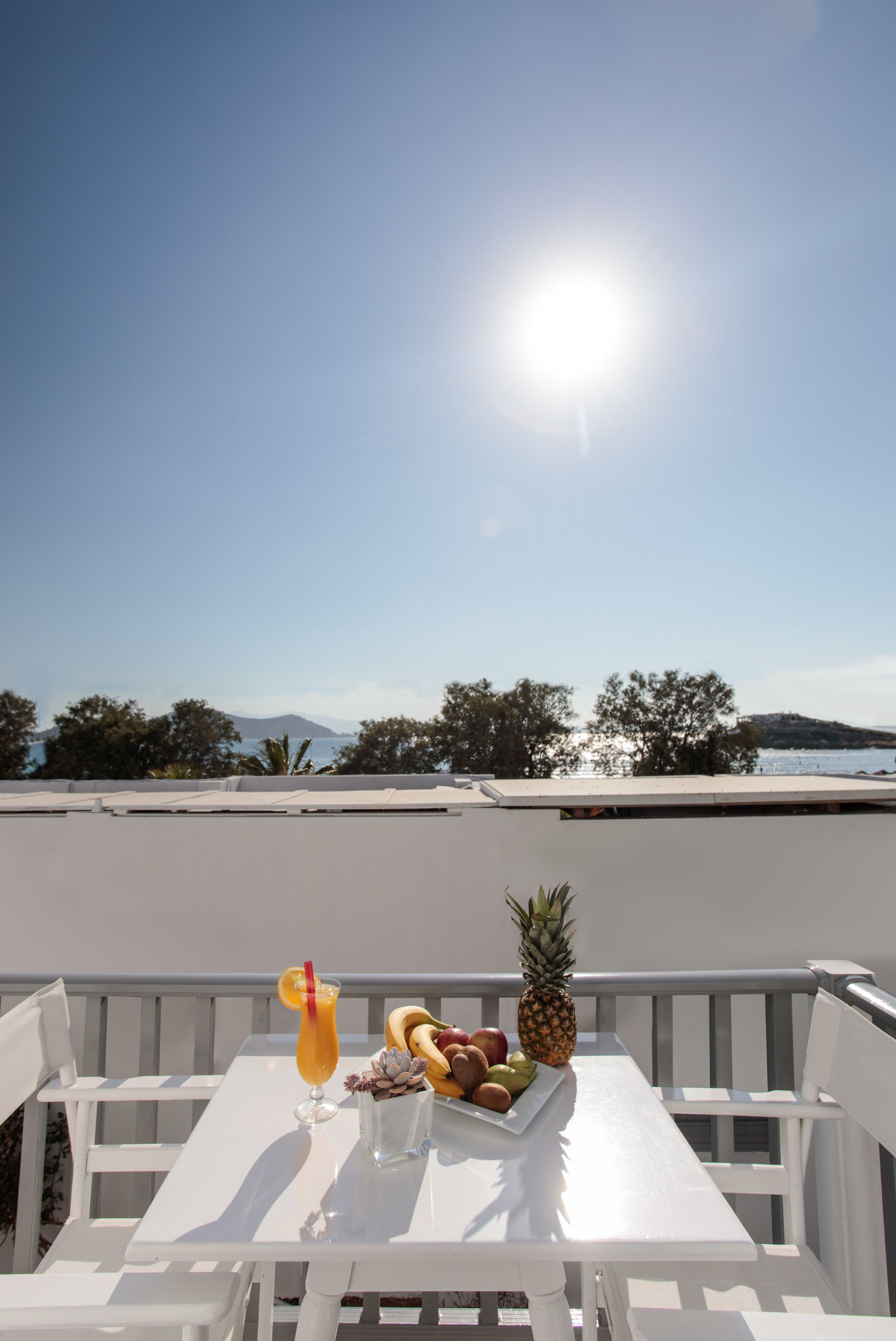 Una mesa blanca con un frutero y una bebida en un balcón con vistas despejadas al mar y árboles.