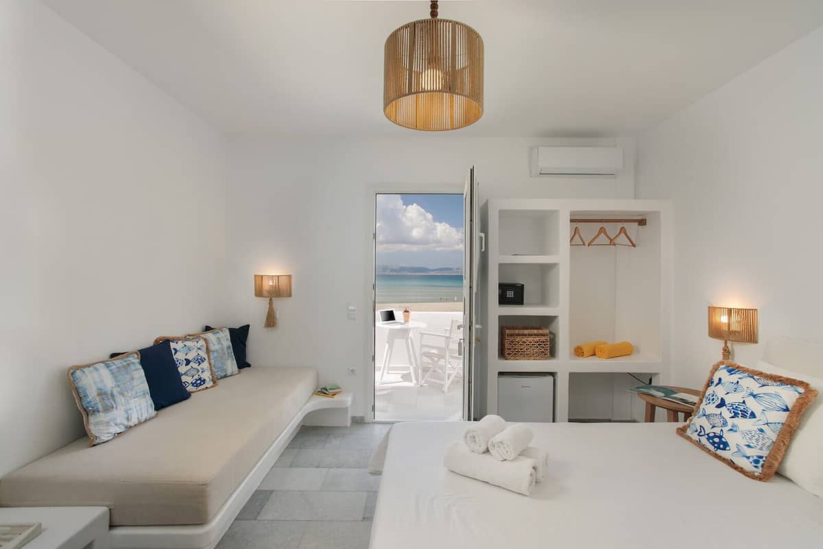 A hotel room with a bed, a seating area with cushions, a wardrobe, and a view of the beach.