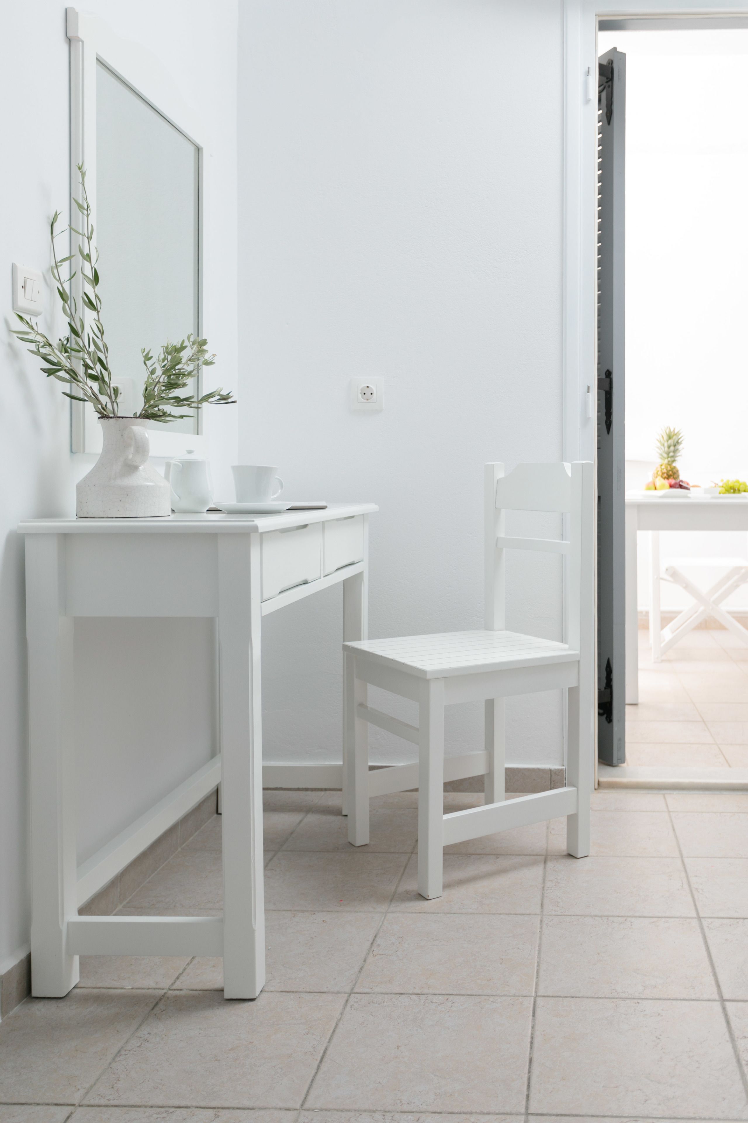 Un baño blanco con espejo, lavamanos, una silla y una mesa con frutas.