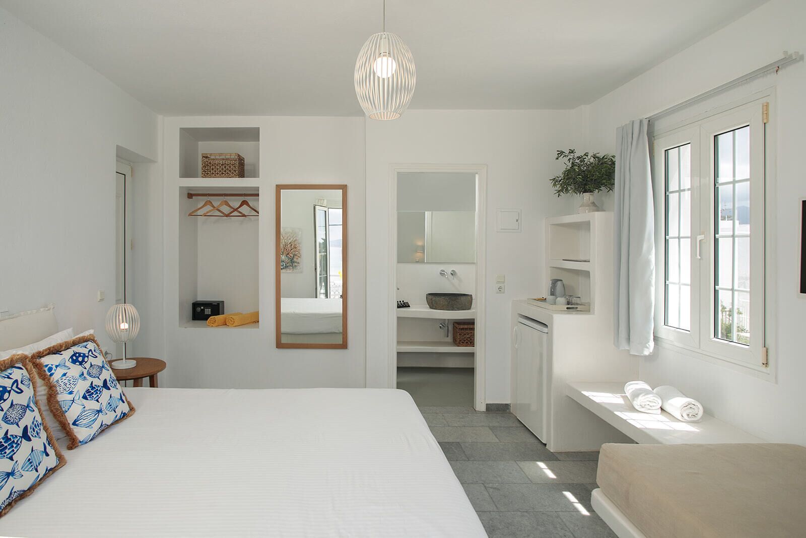 A modern bedroom with a large bed, a bedside table, a mirror, a small desk, a bathroom with a sink and mirror, and a window with curtains.