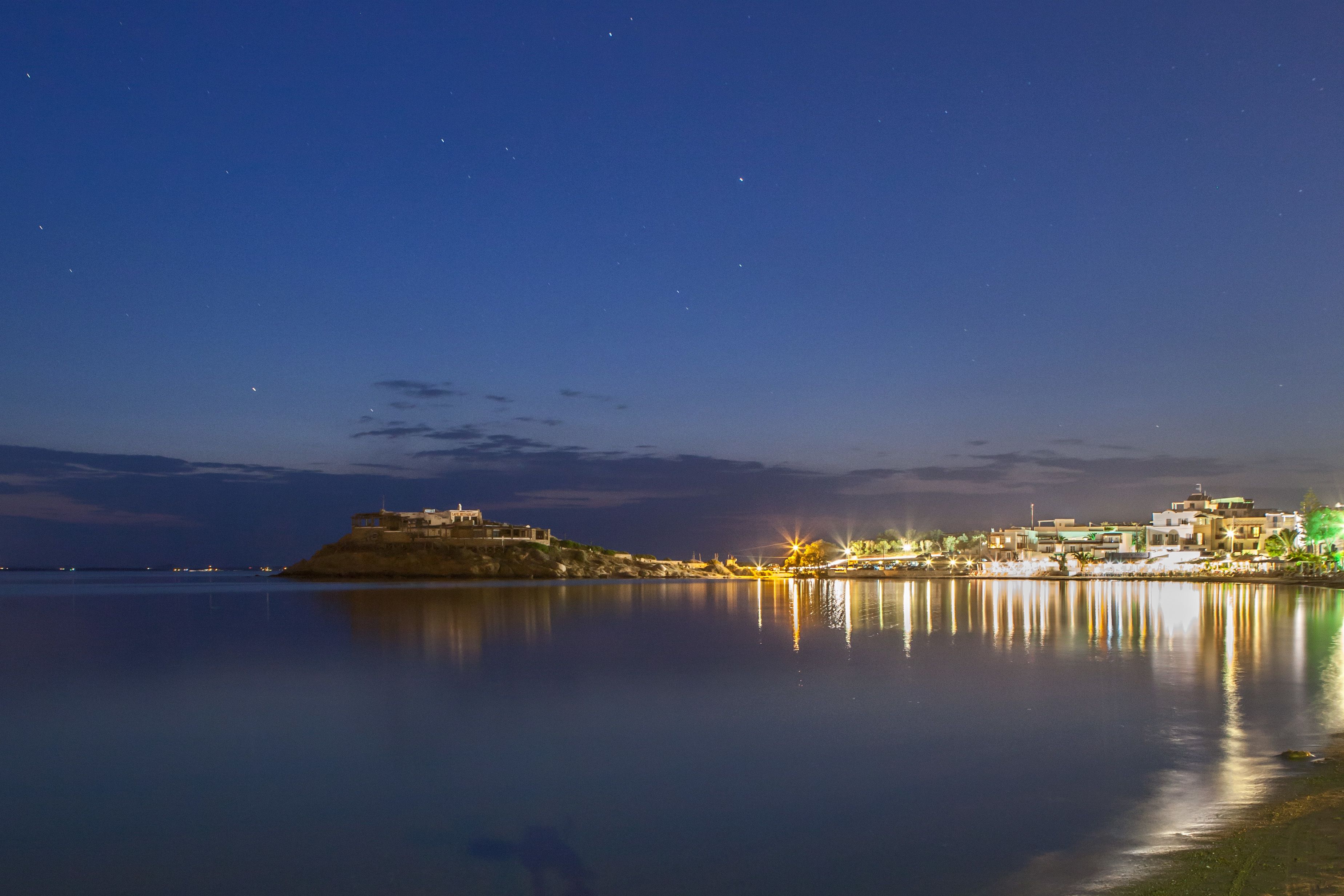 A calm waterfront at night with buildings and a hill in the background.