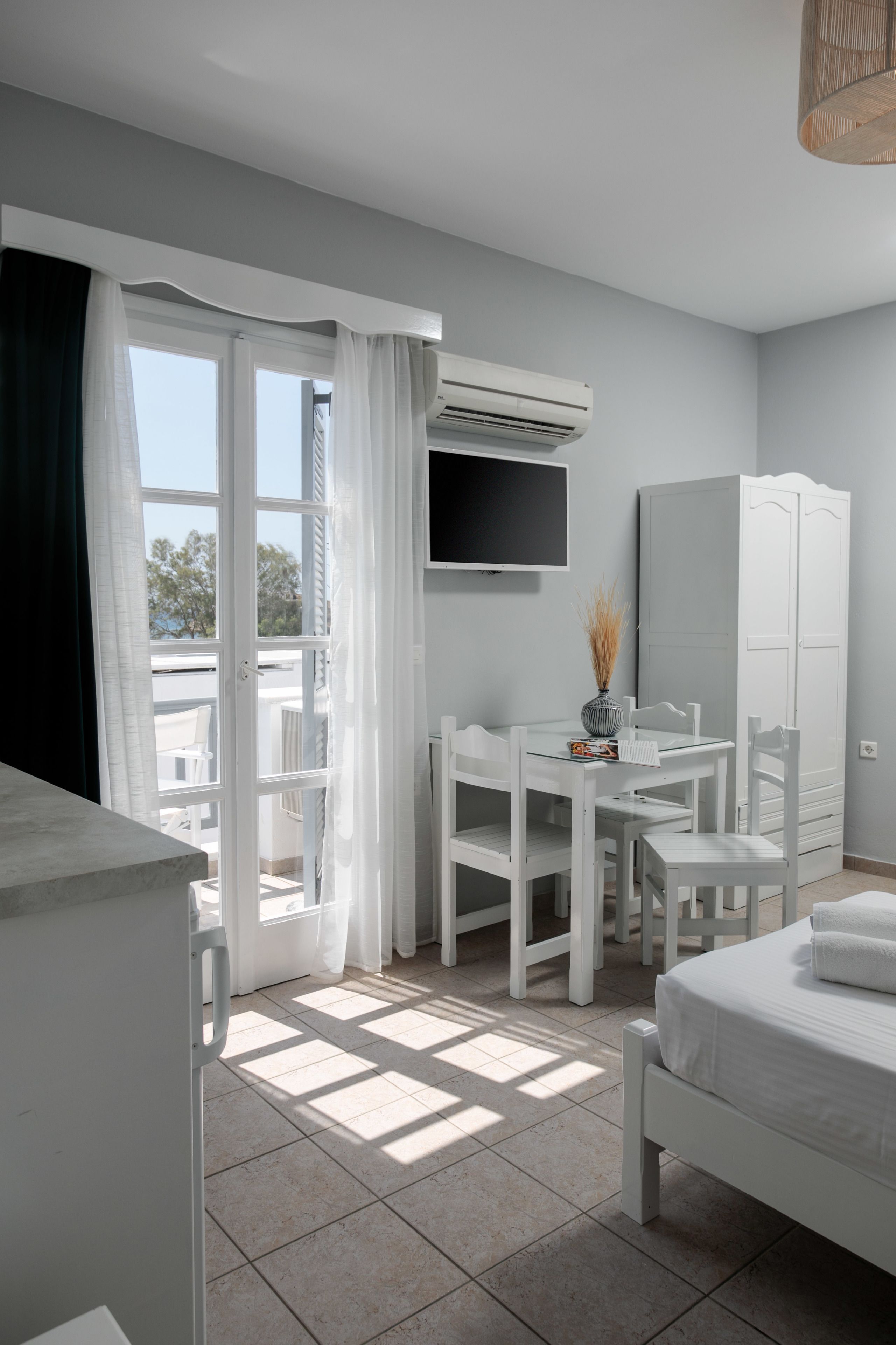 A hotel room with a balcony, a bed, a desk, a chair, a wardrobe, and an air conditioning unit.