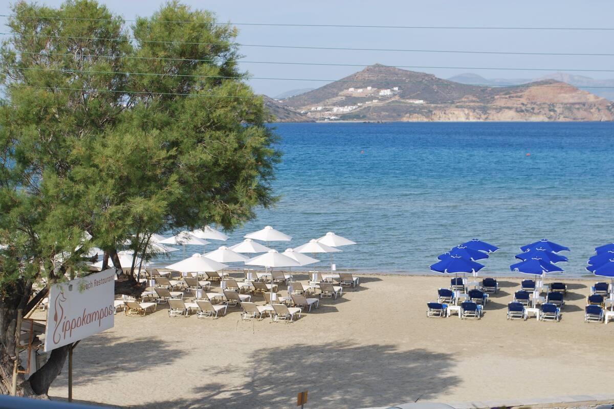 A beach with rows of sun loungers and umbrellas, a sign for 'Eipakampas', and a mountainous backdrop.