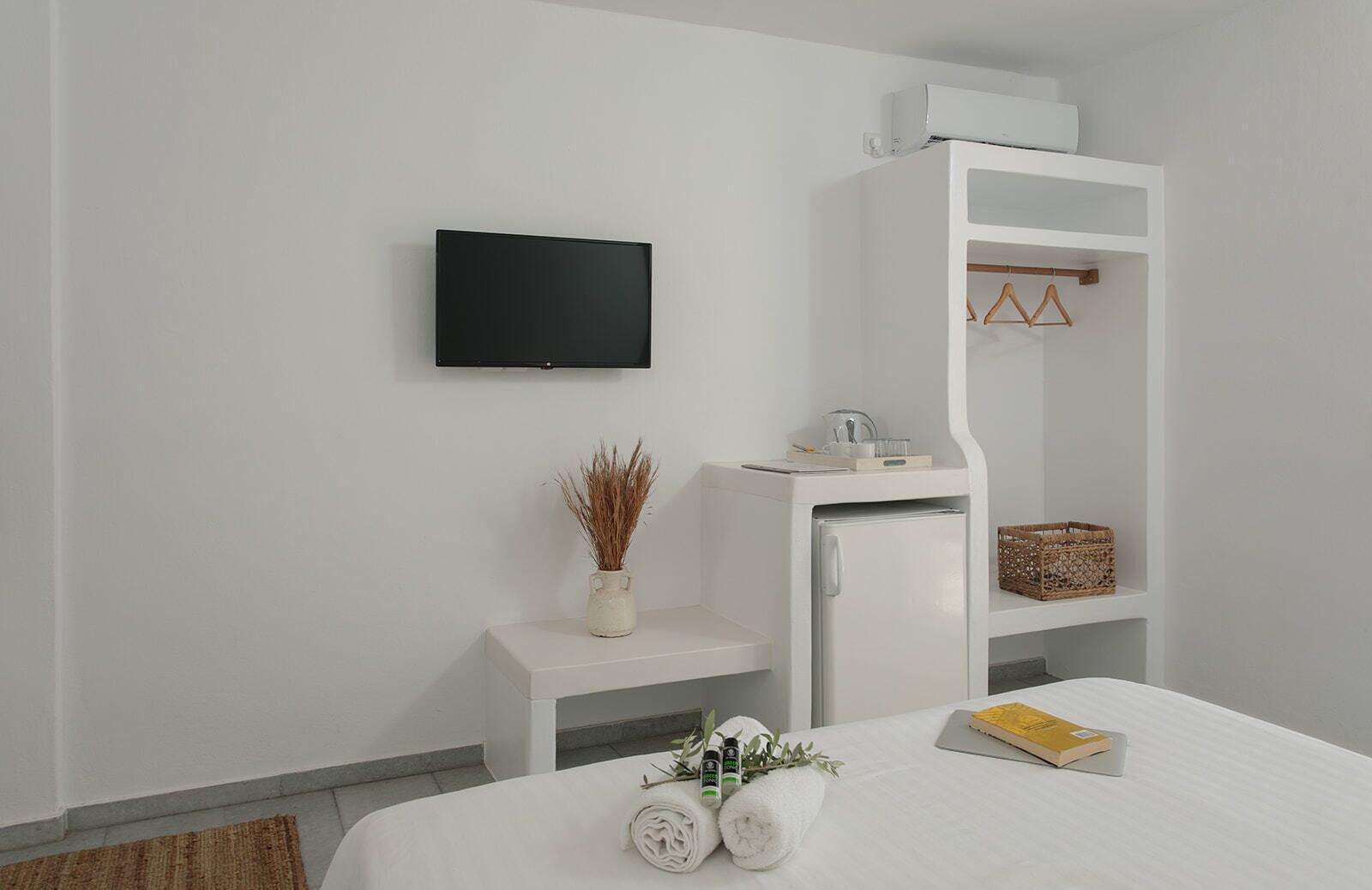 A modern hotel room with a bed, a small white desk, a television, a wardrobe, and a shelf with baskets.