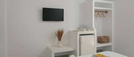 A modern hotel room with a bed, a small white desk, a television, a wardrobe, and a shelf with baskets.