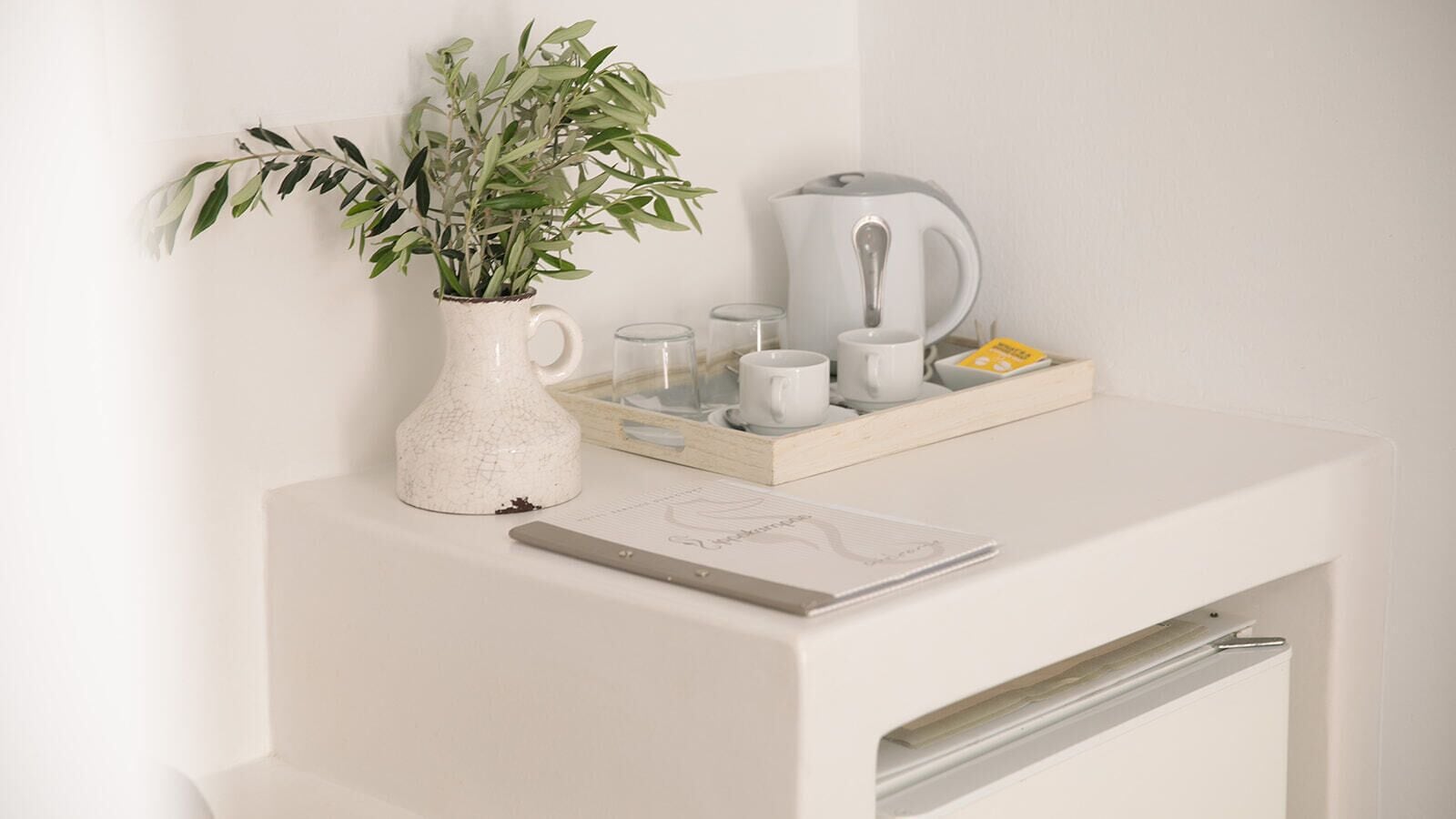 A white desk with a white vase containing green leaves, a kettle, a tray with cups, and a notebook.