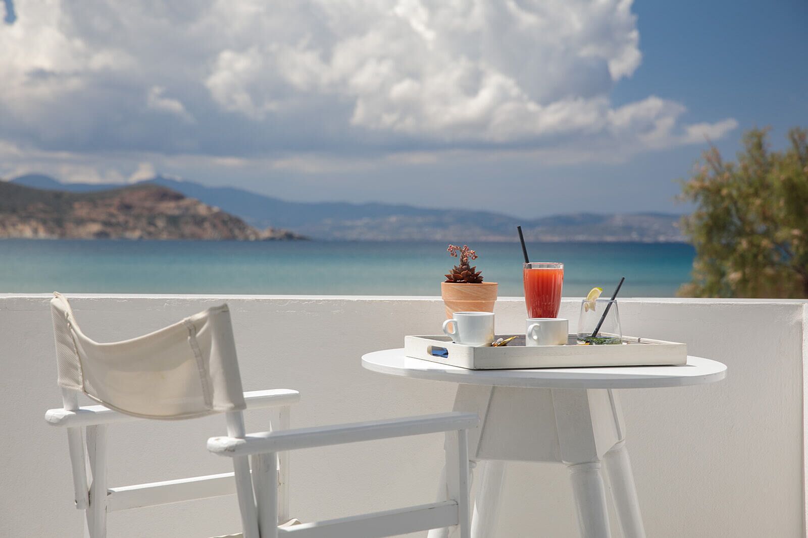 Una silla y una mesa con una bebida y una planta pequeña en un balcón con vistas a la costa.