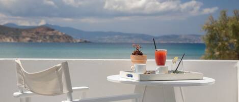 A white chair and table with a drink and a small plant on a balcony overlooking a coastal view.