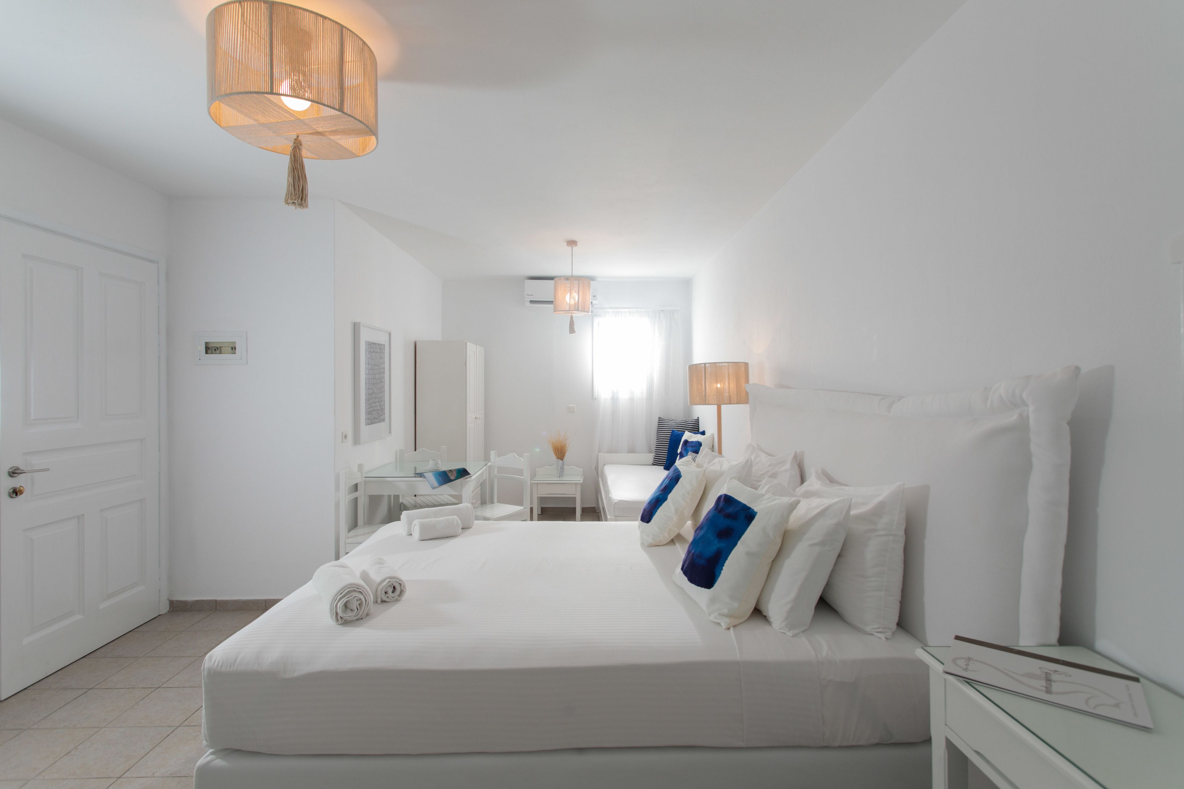 A hotel room with a large bed, a desk, a chair, and a small table. There is a door on the left side of the room.
