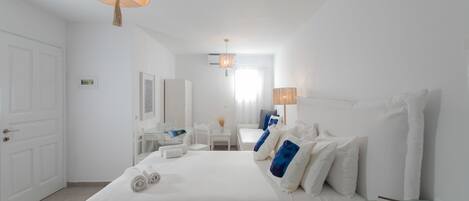 A hotel room with a large bed, a desk, a chair, and a small table. There is a door on the left side of the room.