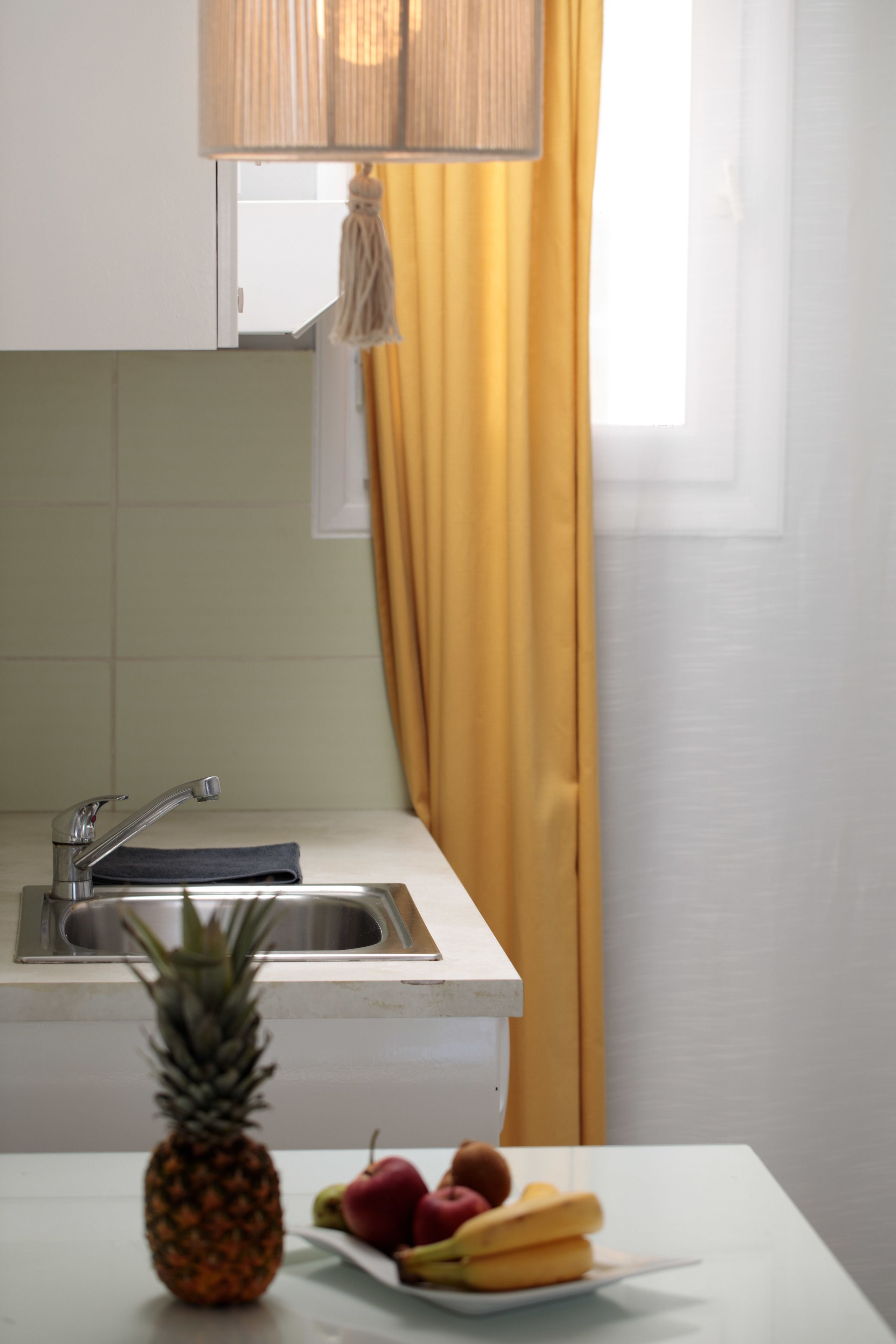 A kitchen with a sink, a window with yellow curtains, and a table with fruits.