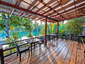Eine Holzterrasse mit Tischen und Stühlen, die auf ein Gewässer mit üppiger Vegetation blickt.