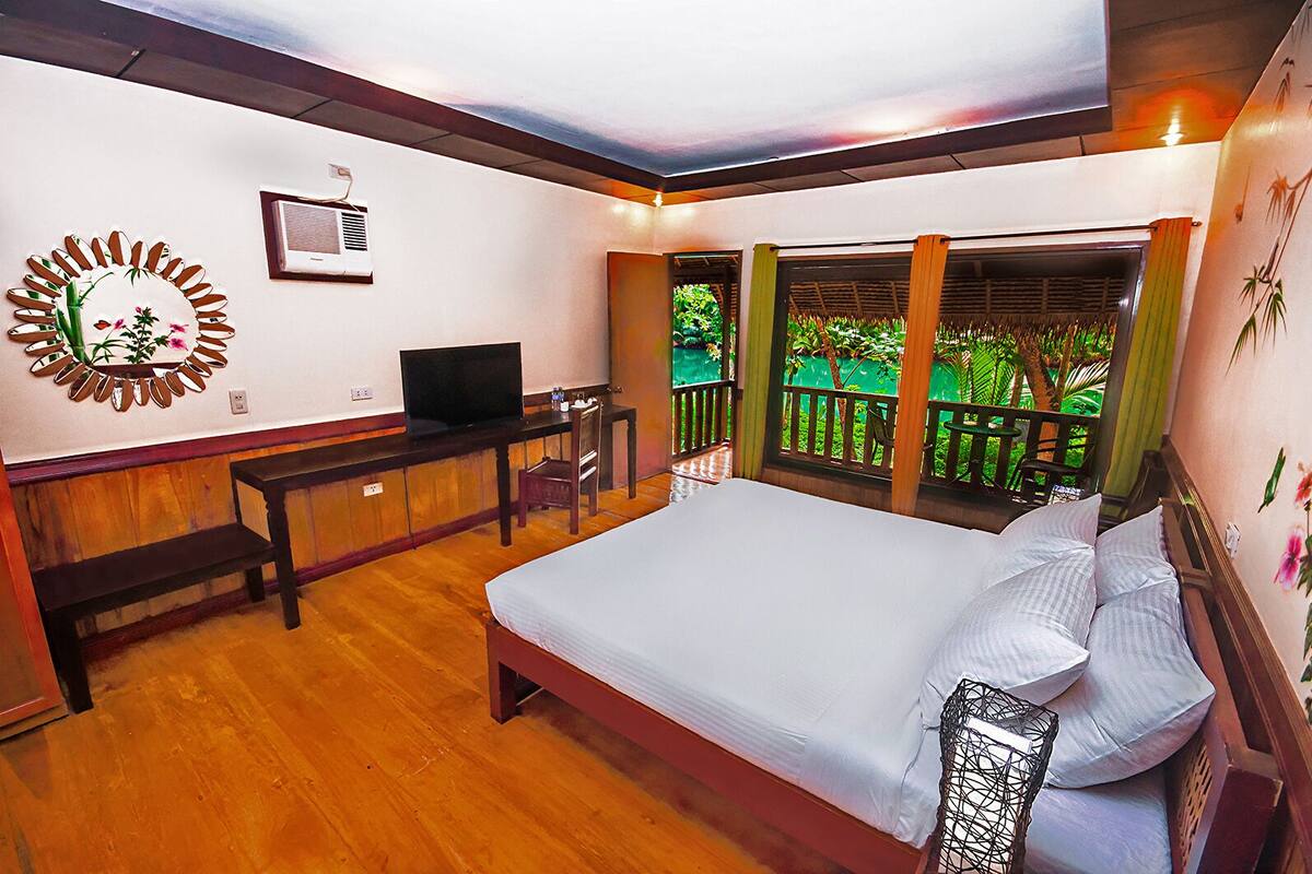 A bedroom with a bed, a desk, a TV, and a view of greenery outside.