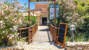A wooden pathway leading to a bar with a signboard displaying the name and a menu. - Piccolo Pevero (Arzachena)