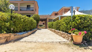 A pathway leading to a building with a tiled roof and a balcony. There is a potted plant with flowers on the right. - Piccolo Pevero (Arzachena)