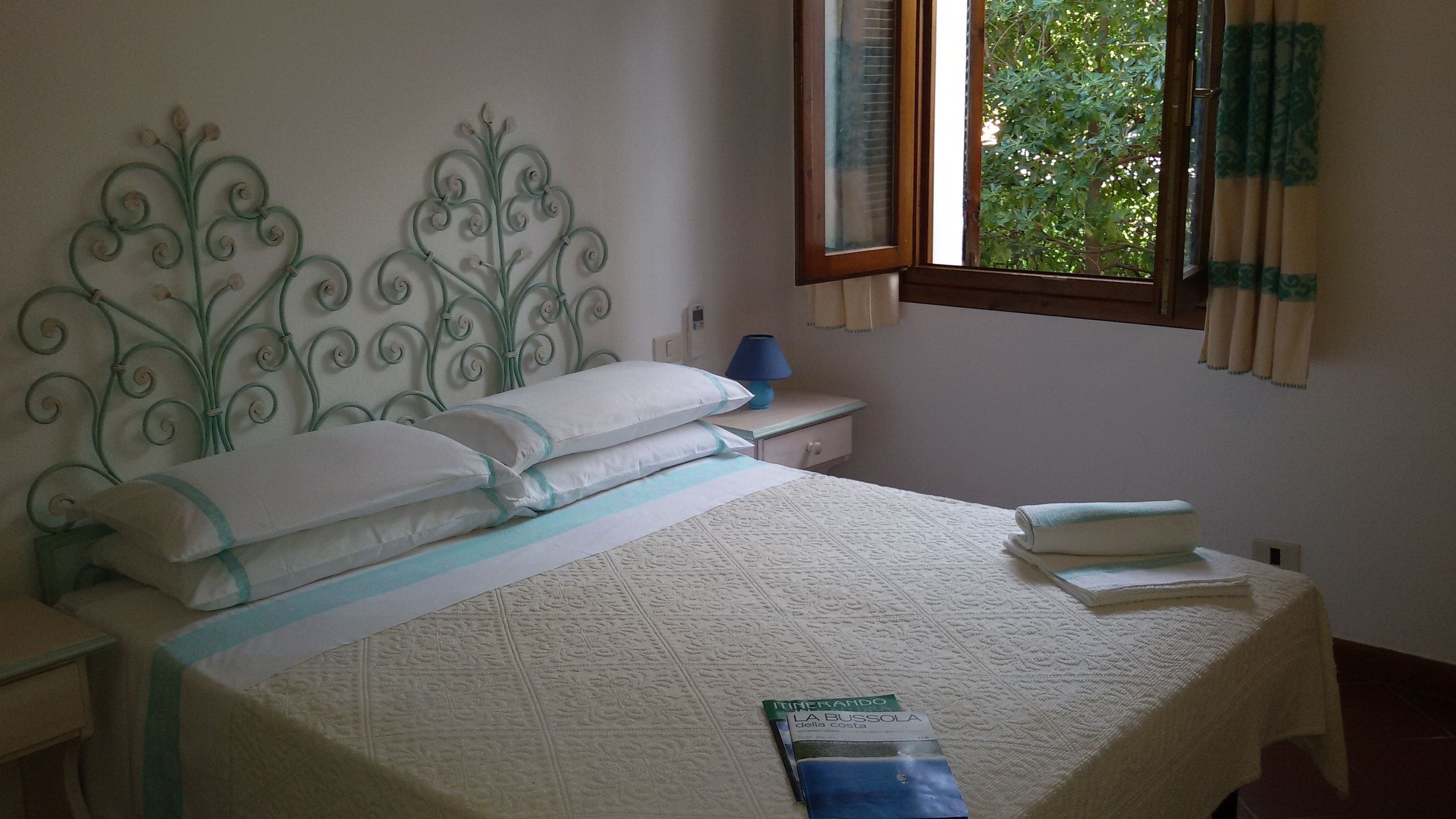 Un letto rifatto con cura, con una coperta bianca e blu, una testiera in metallo decorativa, una lampada da comodino e una finestra con tende.