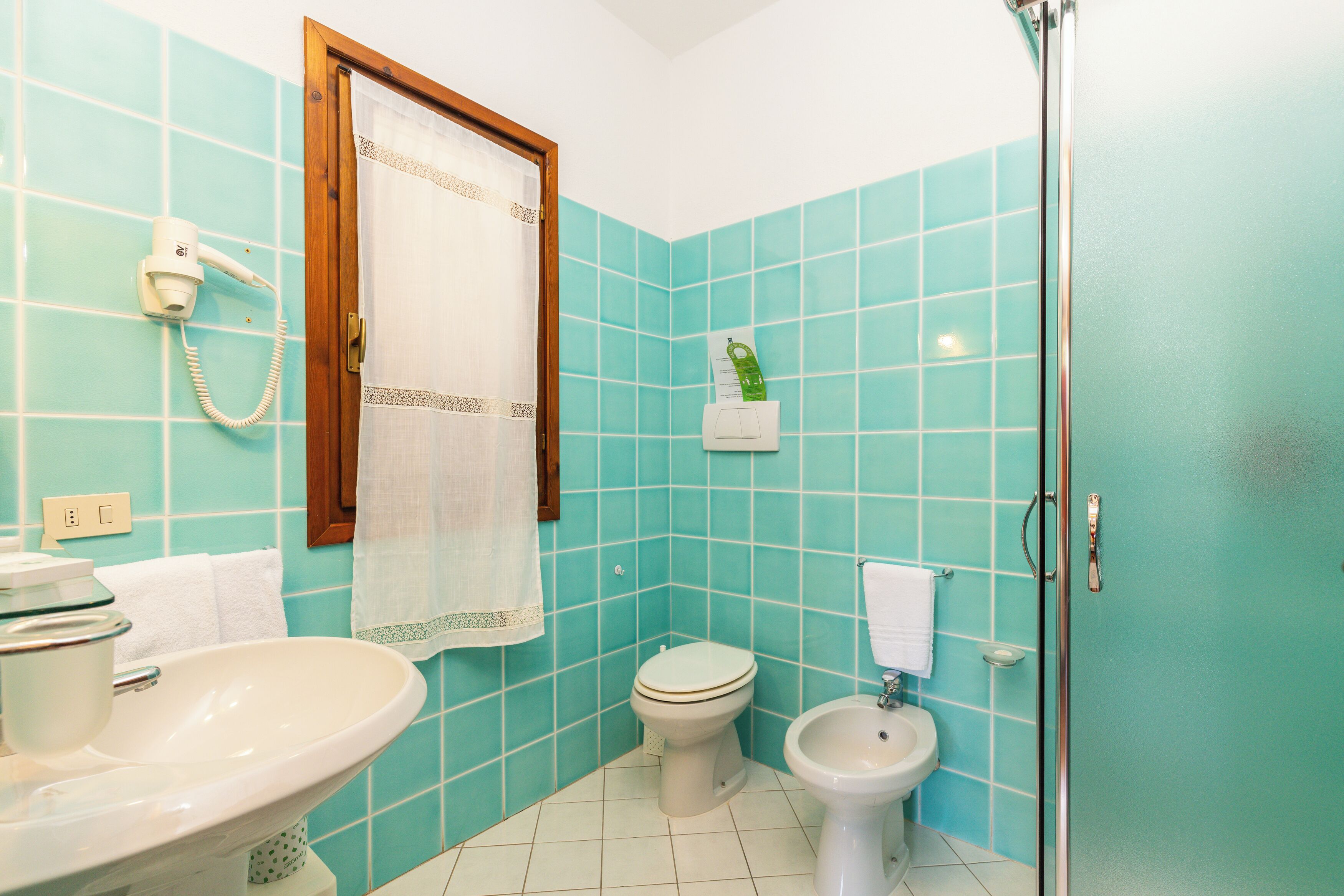 A bathroom with turquoise tiles, a white sink, a toilet, a bidet, a shower with a glass door, and a hairdryer on the wall.