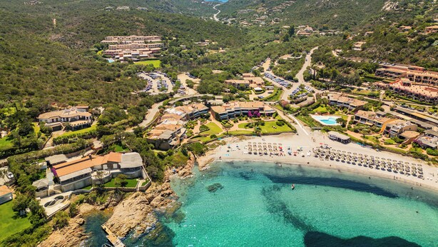 An aerial view of a coastal resort with multiple buildings, a beach with umbrellas, and clear turquoise waters. - Piccolo Pevero (Arzachena)