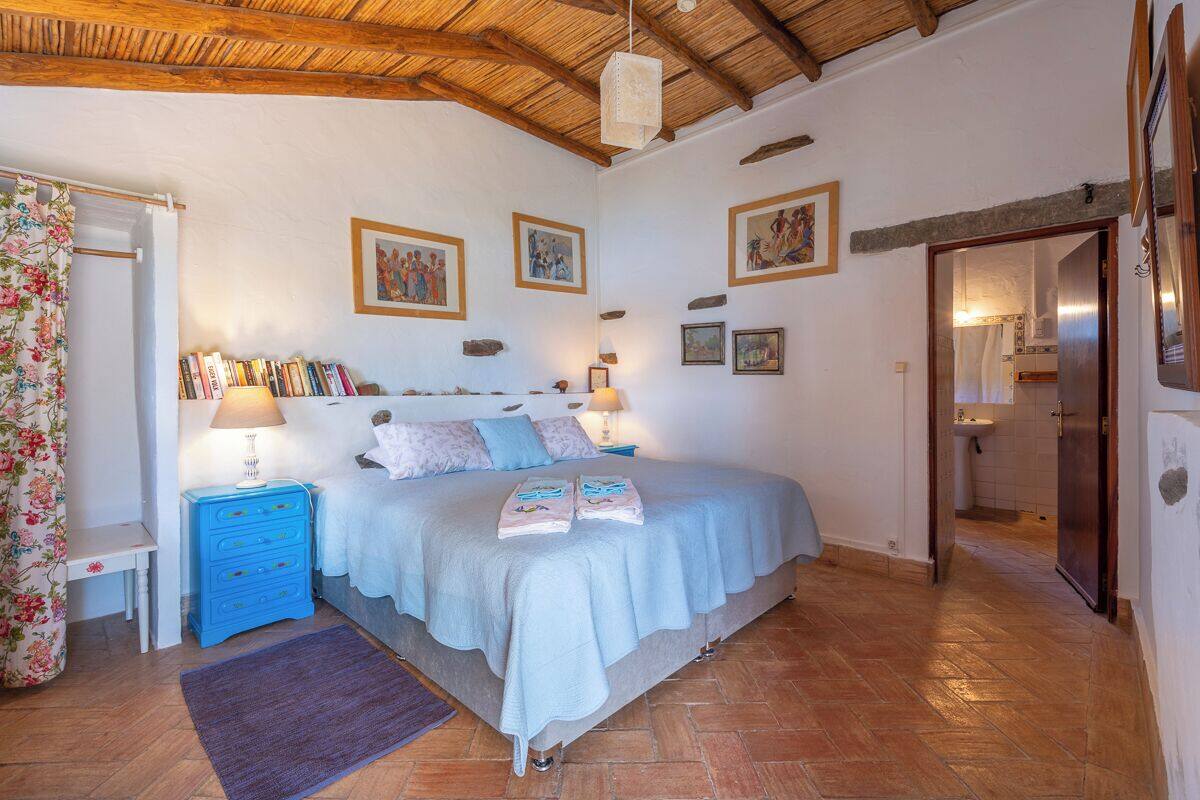 A bedroom with a bed, bedside tables, a nightstand, a wooden ceiling, and a bathroom visible through an open door.