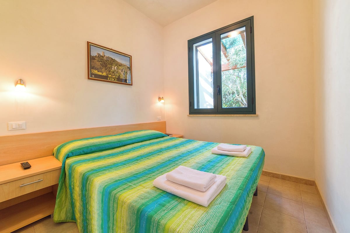 A bedroom with a bed, a nightstand, a wall-mounted light, a window, and a framed picture.
