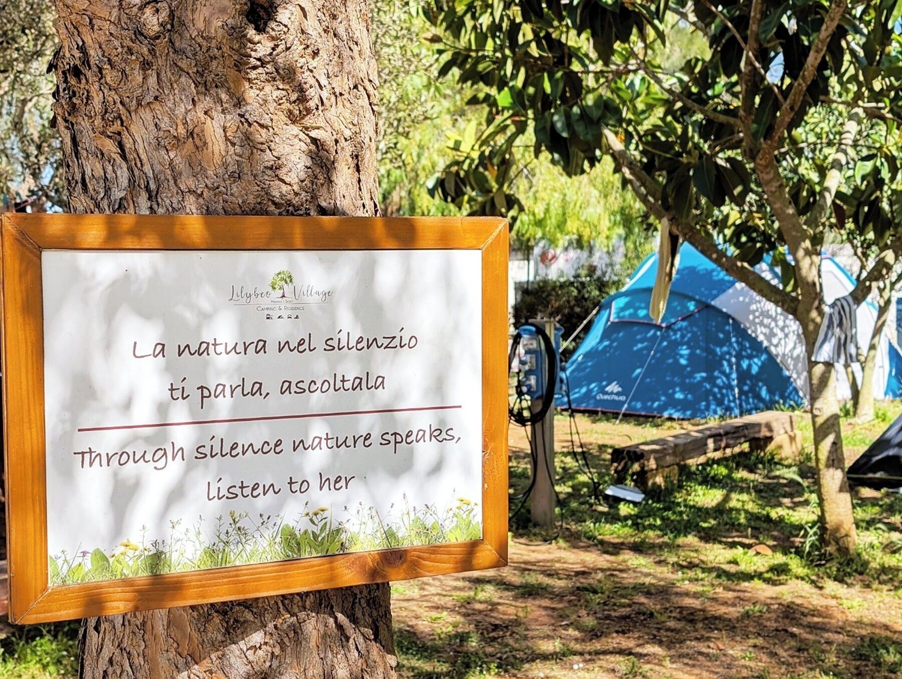 Un cartello con un messaggio sulla voce della natura nel silenzio, posizionato su un ceppo in un campeggio con tende sullo sfondo.