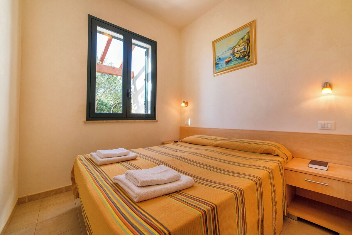 A hotel room with a bed, a nightstand, a wall-mounted lamp, a window with a view, and a painting on the wall.