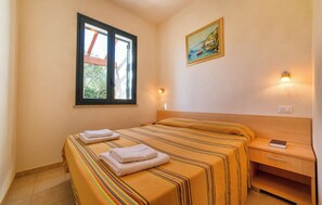 A hotel room with a bed, a nightstand, a wall-mounted lamp, a window with a view, and a painting on the wall. - Lilybeo Village - Camping & Residence (Marsala)