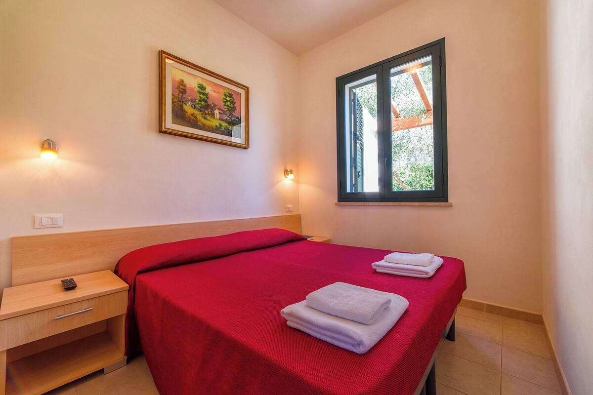 A bedroom with a wooden headboard, a red bedspread, two white towels, a nightstand, a wall-mounted light, a framed painting, and a window with a view of greenery.