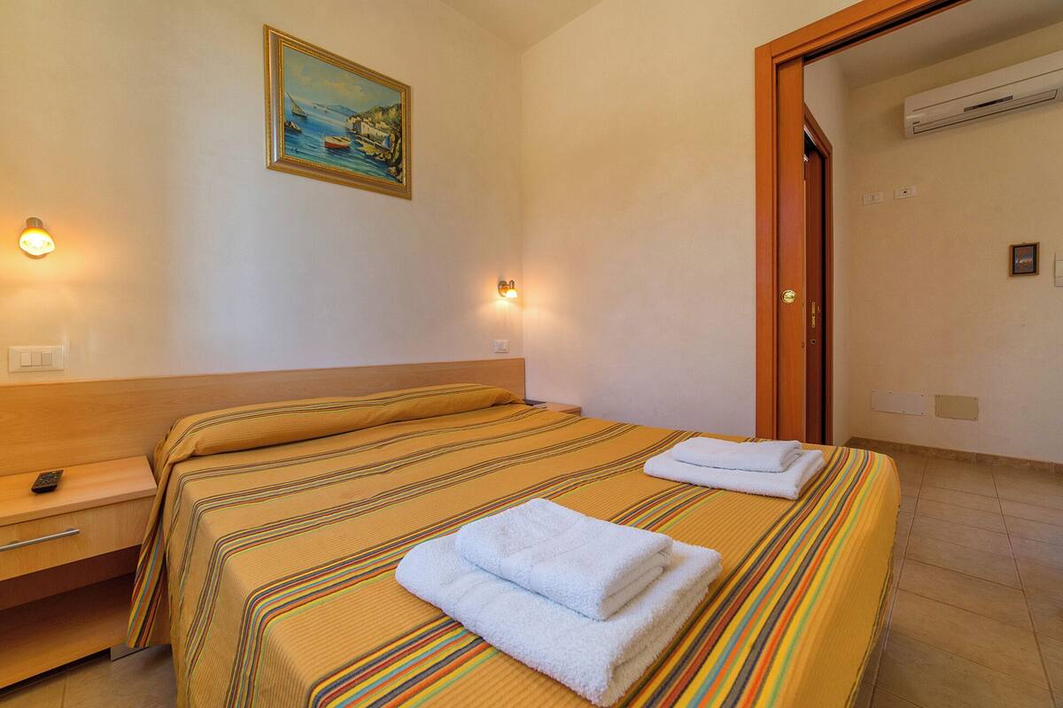A hotel room with a bed, bedside table, nightstand, and a painting on the wall.