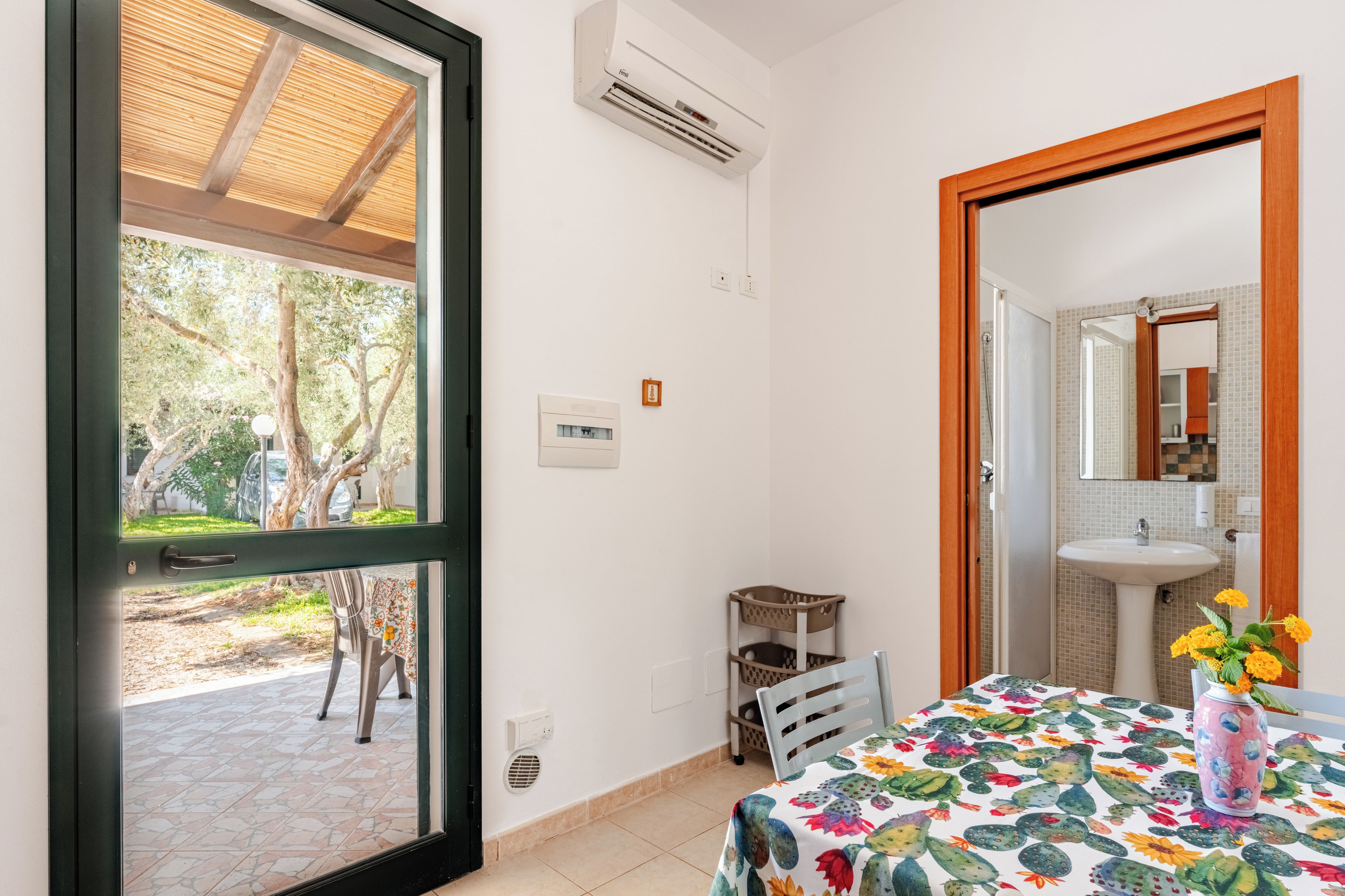 Een kamer met een raam, een airconditioningapparaat, een tafel met een bloemig tafelkleed, een stoel en een vaas met bloemen.