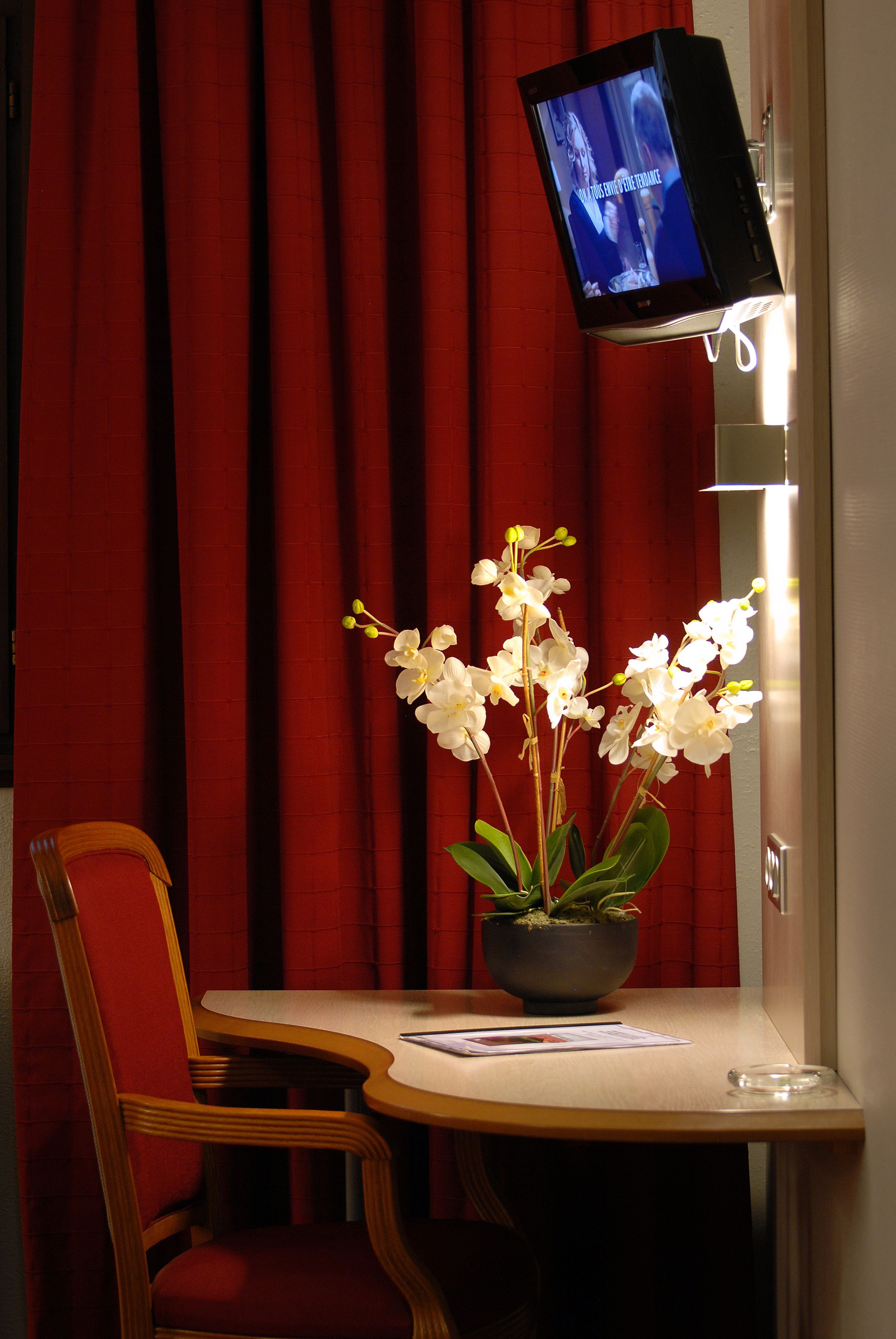 A room with a television mounted on the wall, a wooden chair, a small table with a vase of white orchids, and red curtains.