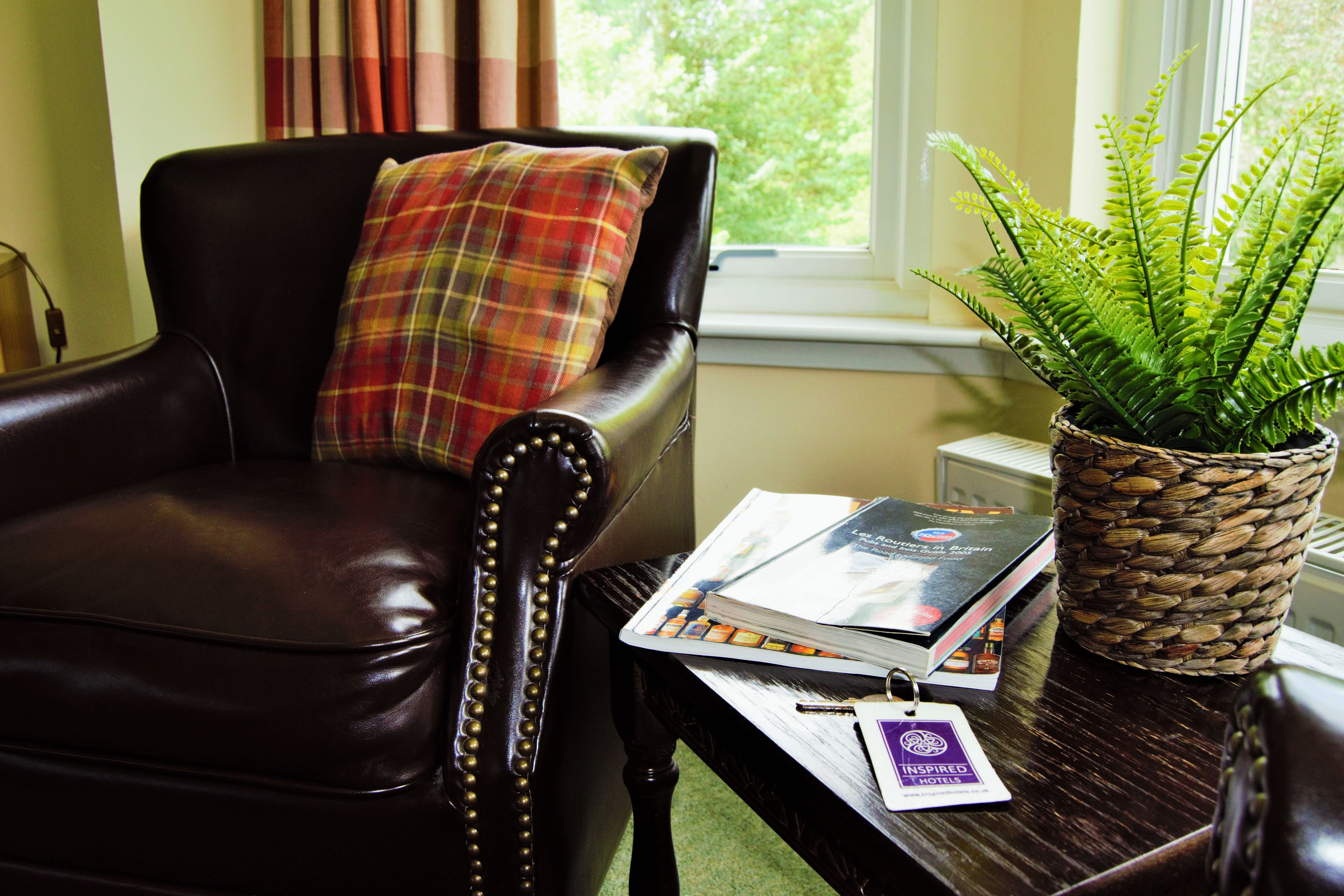 A leather armchair with a plaid pillow, a wooden coffee table with a book and a plant, and a window with a view of trees.