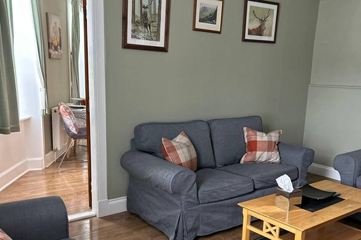 A living room with a grey sofa, a wooden coffee table, and a small rug. There are two framed pictures on the wall and a door leading to another room.