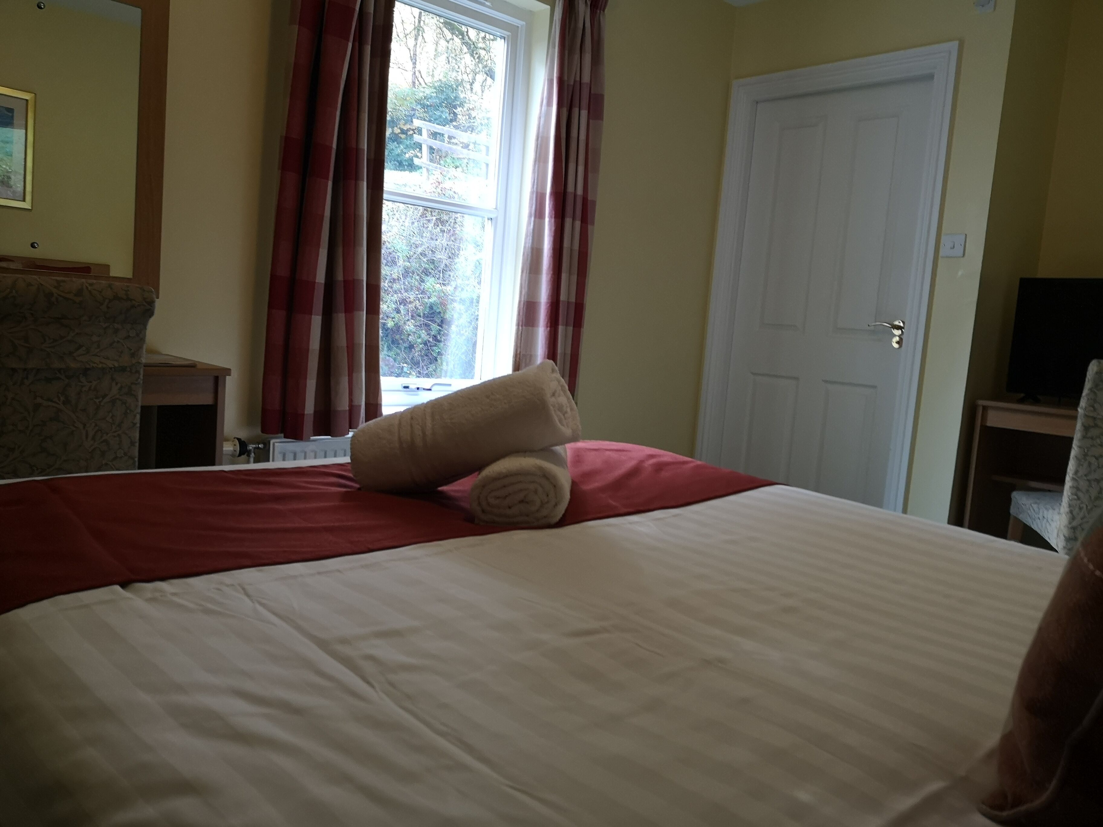 Una cama bien hecha con una colcha roja y blanca, una toalla beige enrollada y una almohada roja. Hay una ventana con cortinas rojas, una puerta blanca y un televisor sobre un mueble a la derecha.