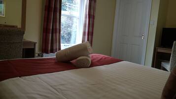Una cama bien hecha con una colcha roja y blanca, una toalla beige enrollada y una almohada roja. Hay una ventana con cortinas rojas, una puerta blanca y un televisor sobre un mueble a la derecha.