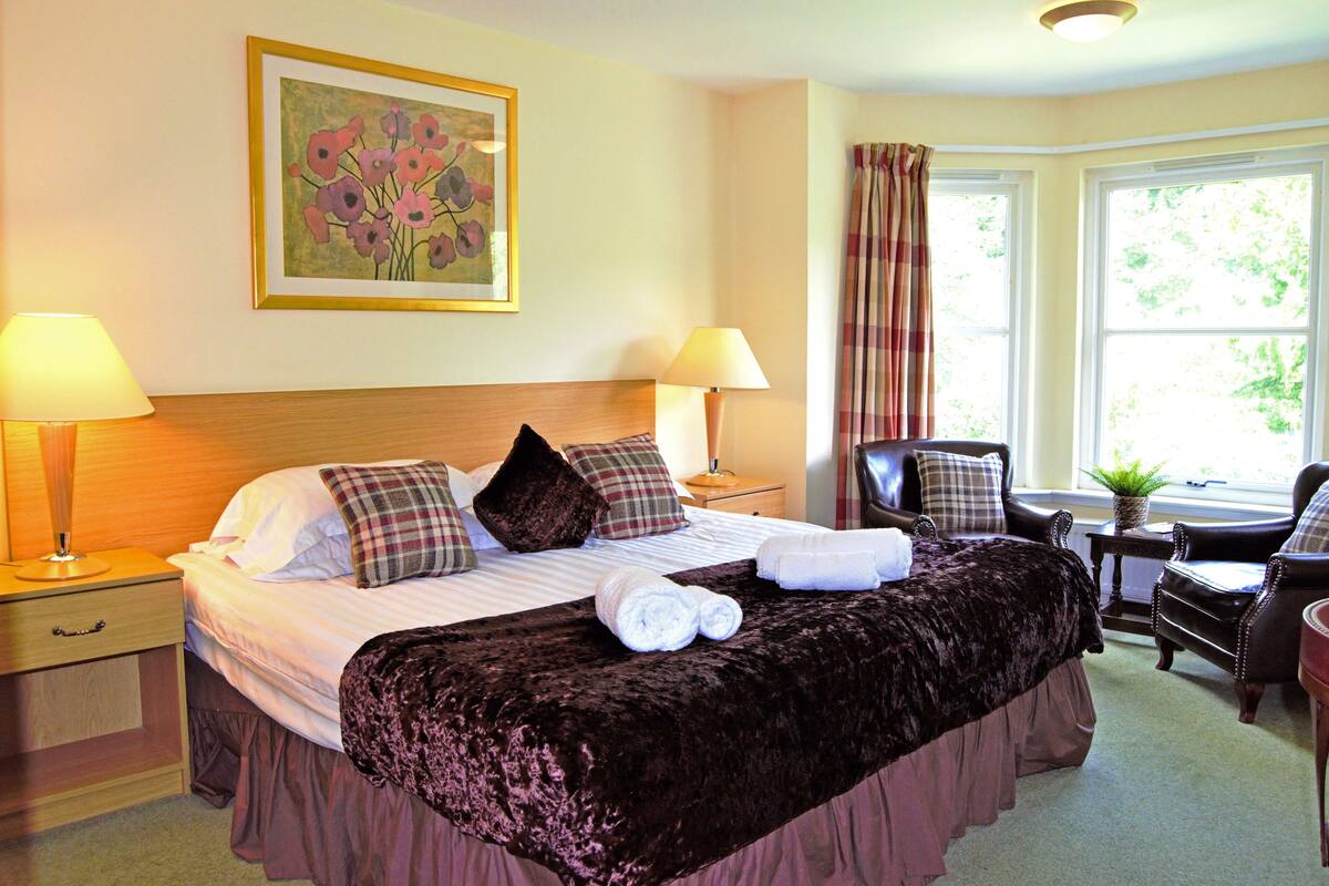 A hotel room with a large bed, two bedside tables with lamps, a desk with a chair, and a window with curtains.