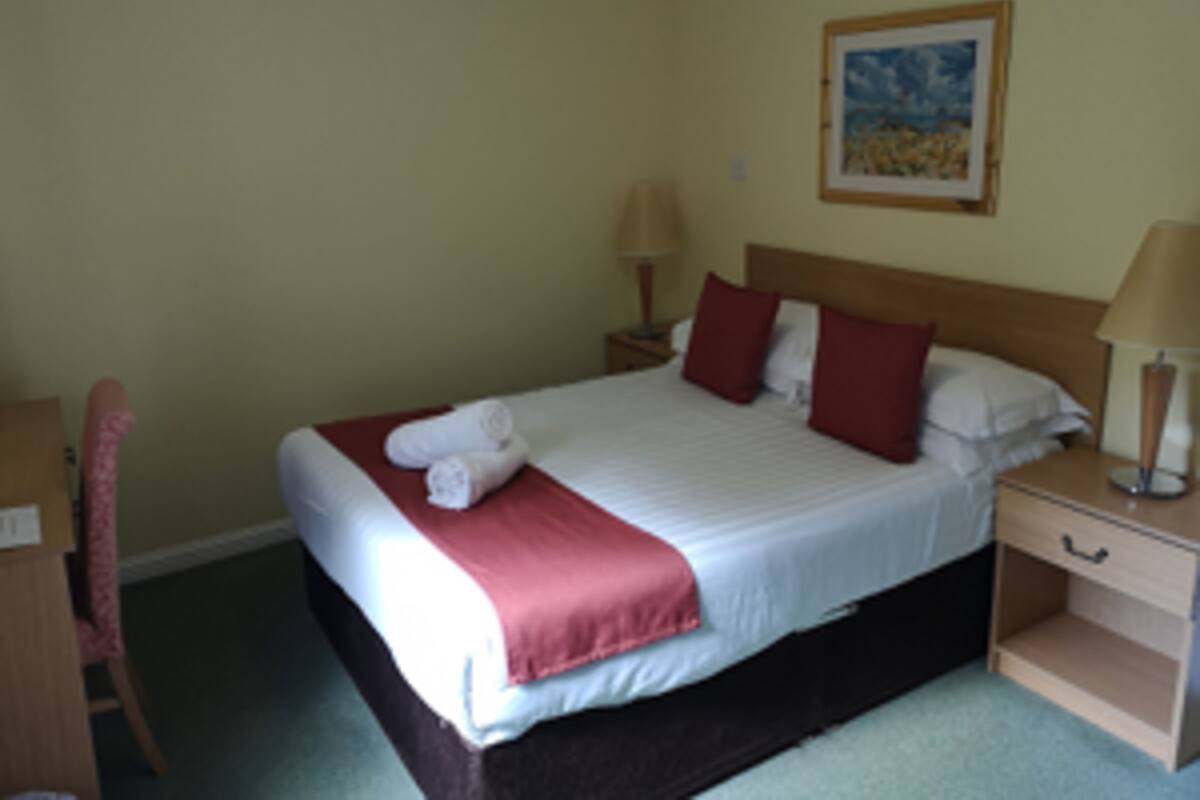 A neatly made bed with a white comforter and red pillows, a wooden nightstand with a lamp, and a framed picture on the wall.