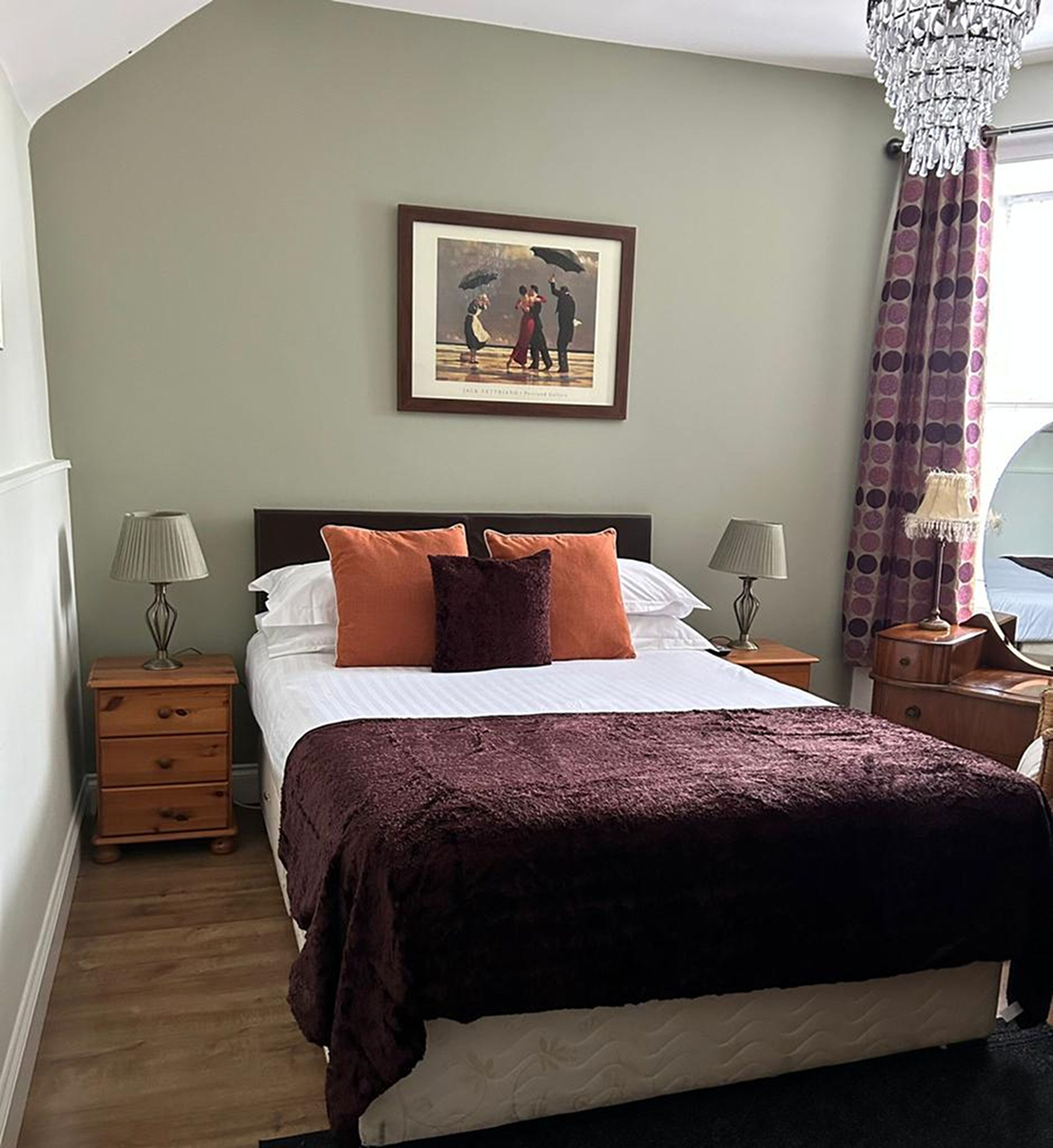 Een slaapkamer met een bed, twee nachtkastjes, een kroonluchter, een ingelijst schilderij aan de muur en een raam met gordijnen.