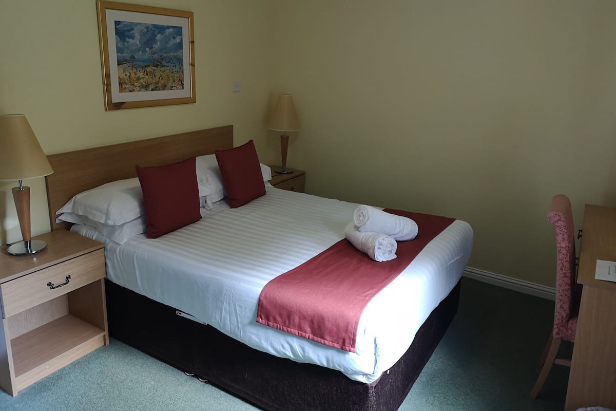A neatly made bed with white and red bedding, two bedside lamps, a wooden nightstand, a small desk, and a framed picture on the wall.