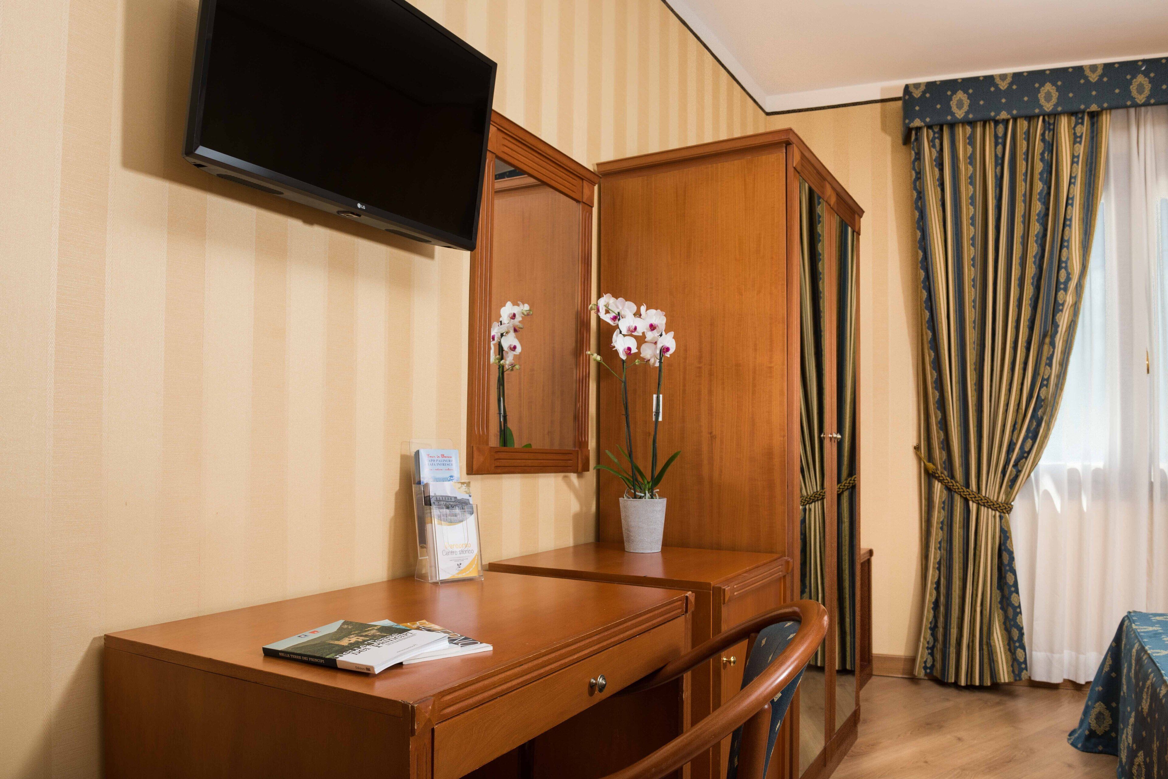 Une chambre d’hôtel avec un bureau en bois, un téléviseur à écran plat fixé au mur, une orchidée en pot et une fenêtre avec des rideaux à rayures.