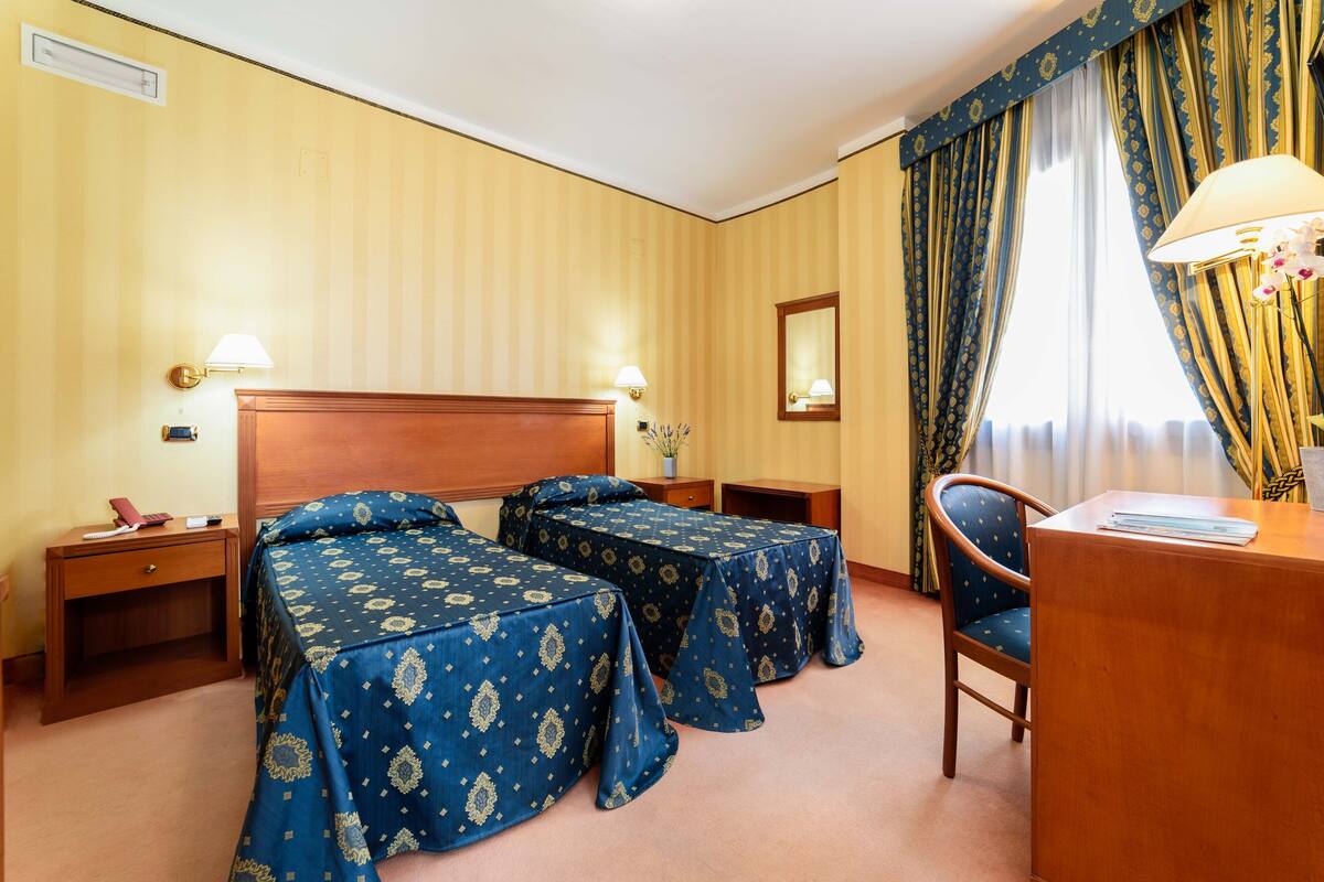 A hotel room with two beds, a desk, a chair, a nightstand, and a lamp.